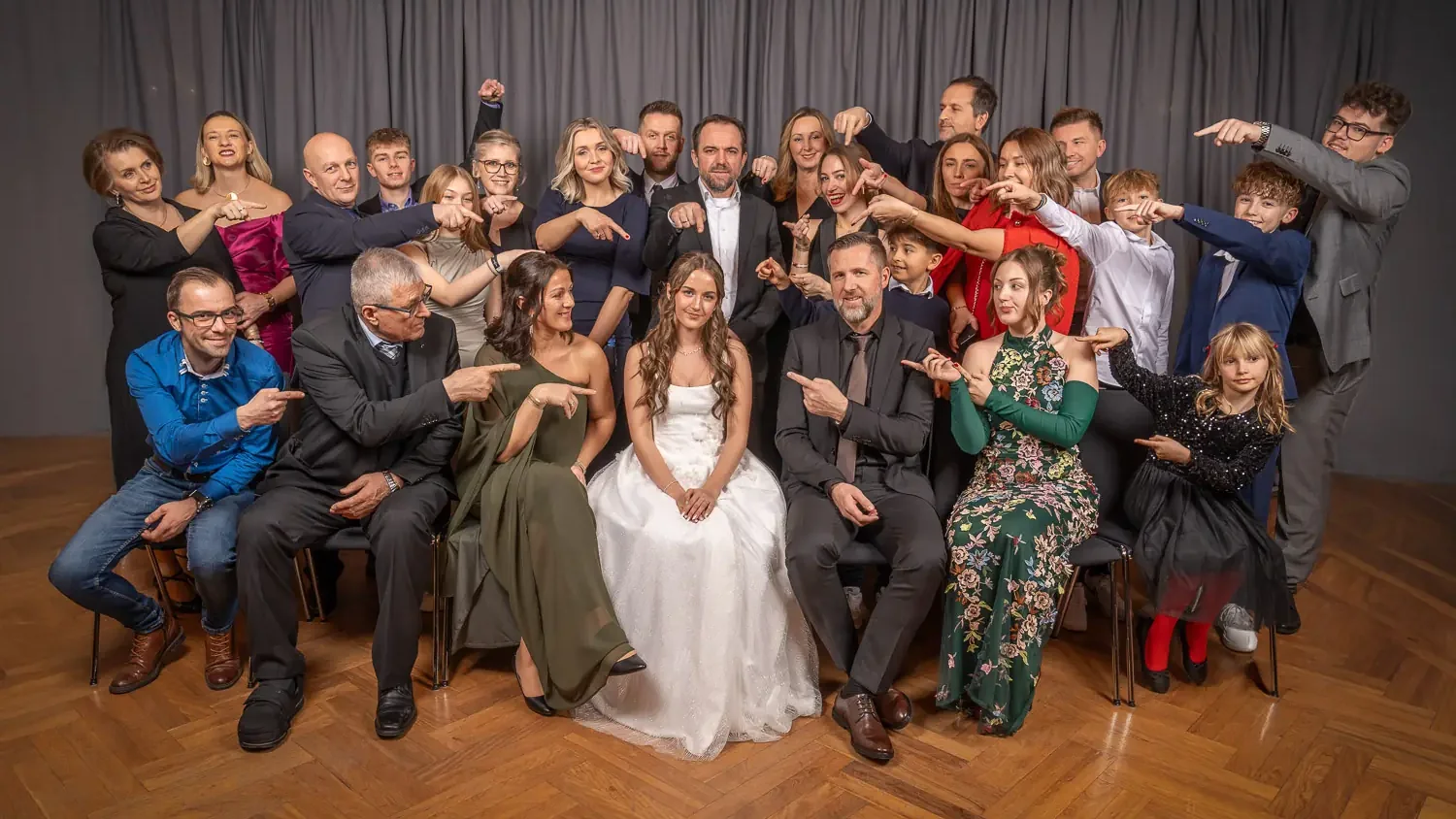 Familienfoto im Ballstudio: Maturantin im weißen Ballkleid, Familie und Freunde zeigen lachend auf sie